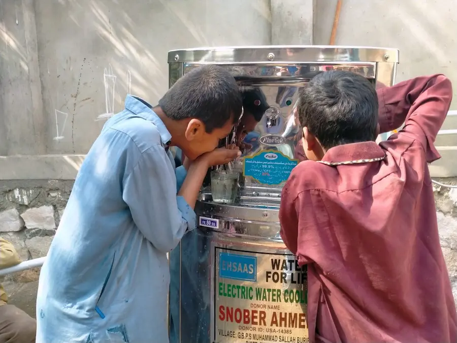 Two boys drinking clean water from an electric water cooler installed under a donor program for safe drinking water by SDO.