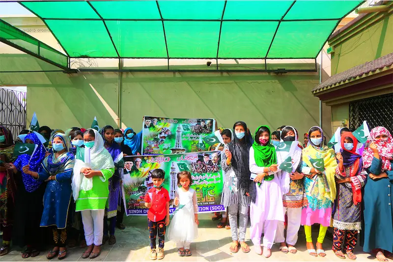 Sindh Development Organization (SDO) NGO event in Karachi, Pakistan, with women and children holding flags and banners, celebrating national pride and sustainability, September 2025.